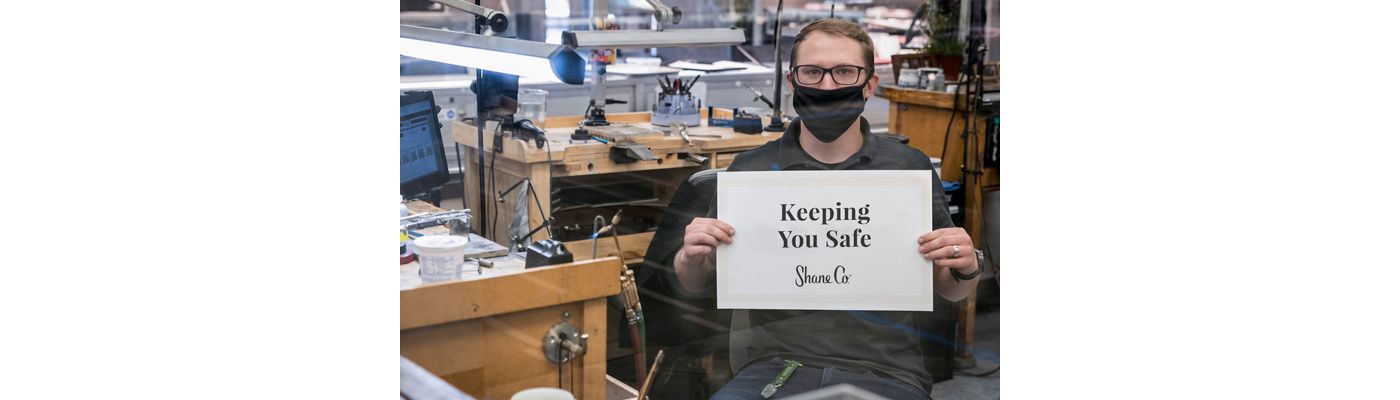A jeweler wearing a face covering and holding a sign that reads keeping you safe